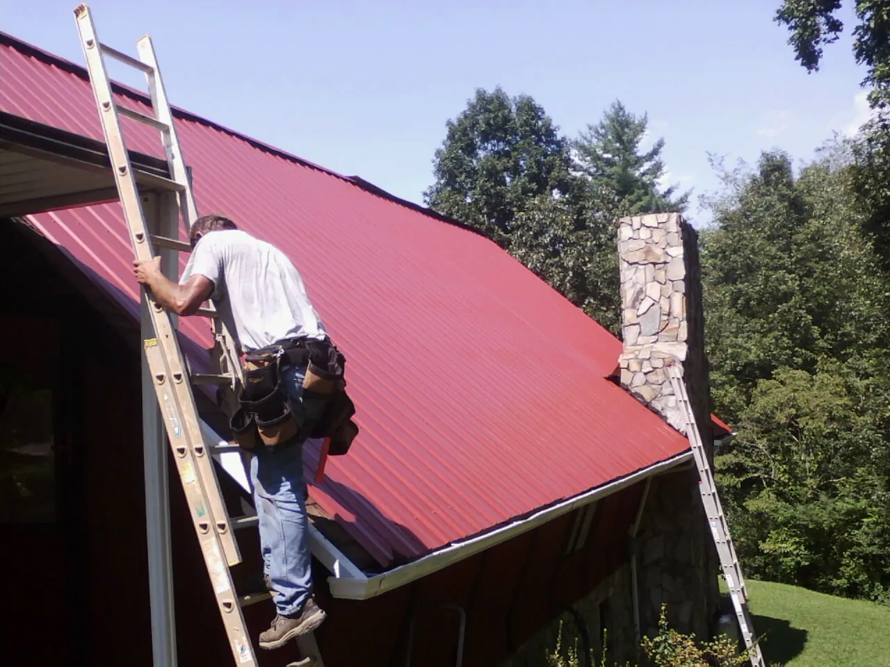 Roofer on a ladder beside a red metal roof and stone chimney — Soffit & Fascia Repair by Roofing Brothers in Wahpeton